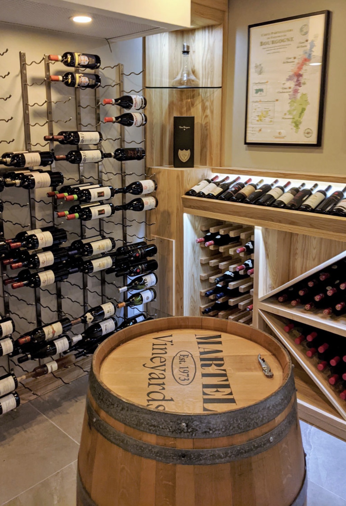 Cave à vin aménagée avec casiers métalliques, rayonnages en bois clair éclairés et tonneau servant de table de dégustation
