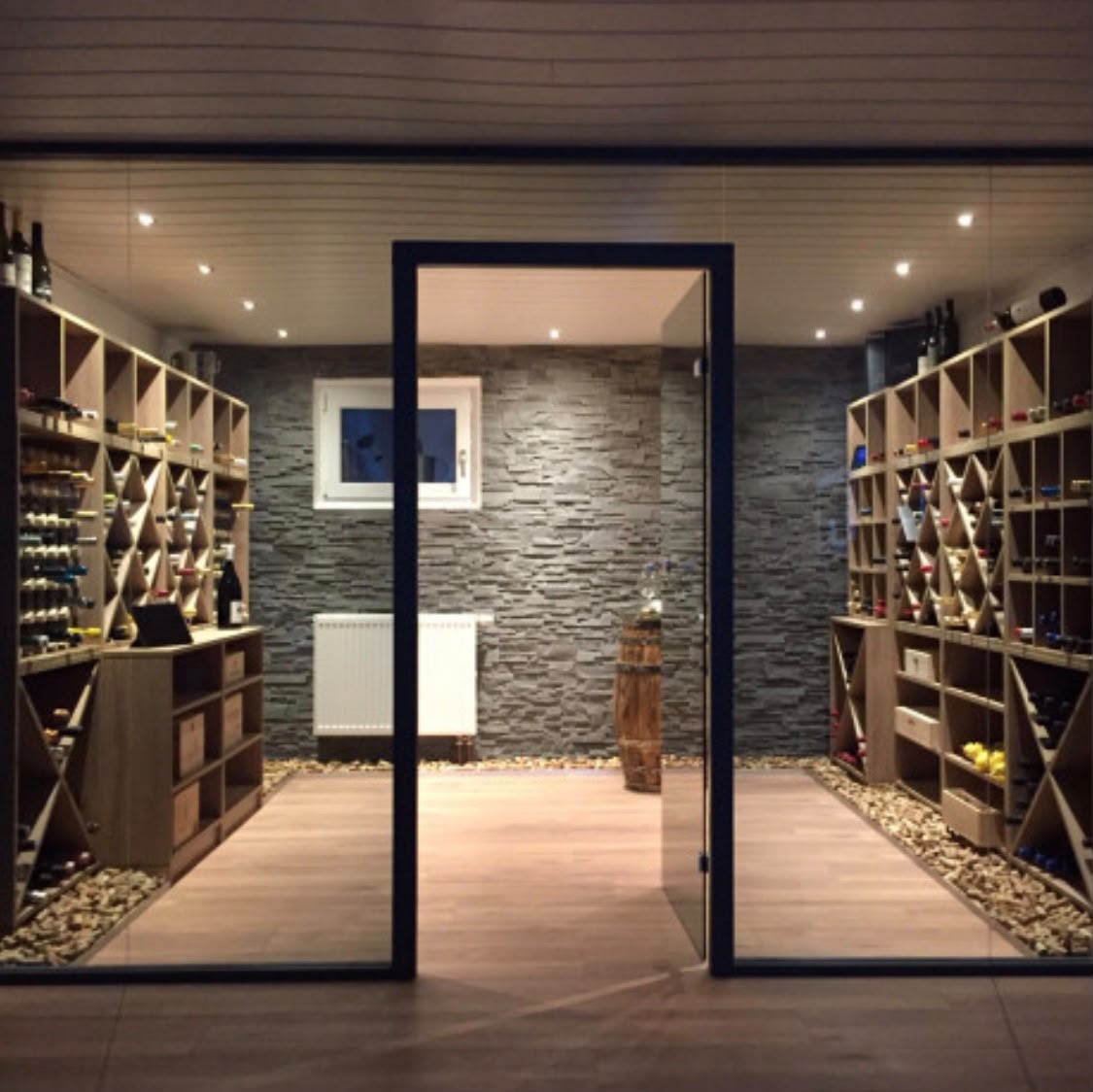 Cave à vin vitrée avec porte en verre, casiers en bois de part et d'autre, mur en pierre et lit de galets au sol
