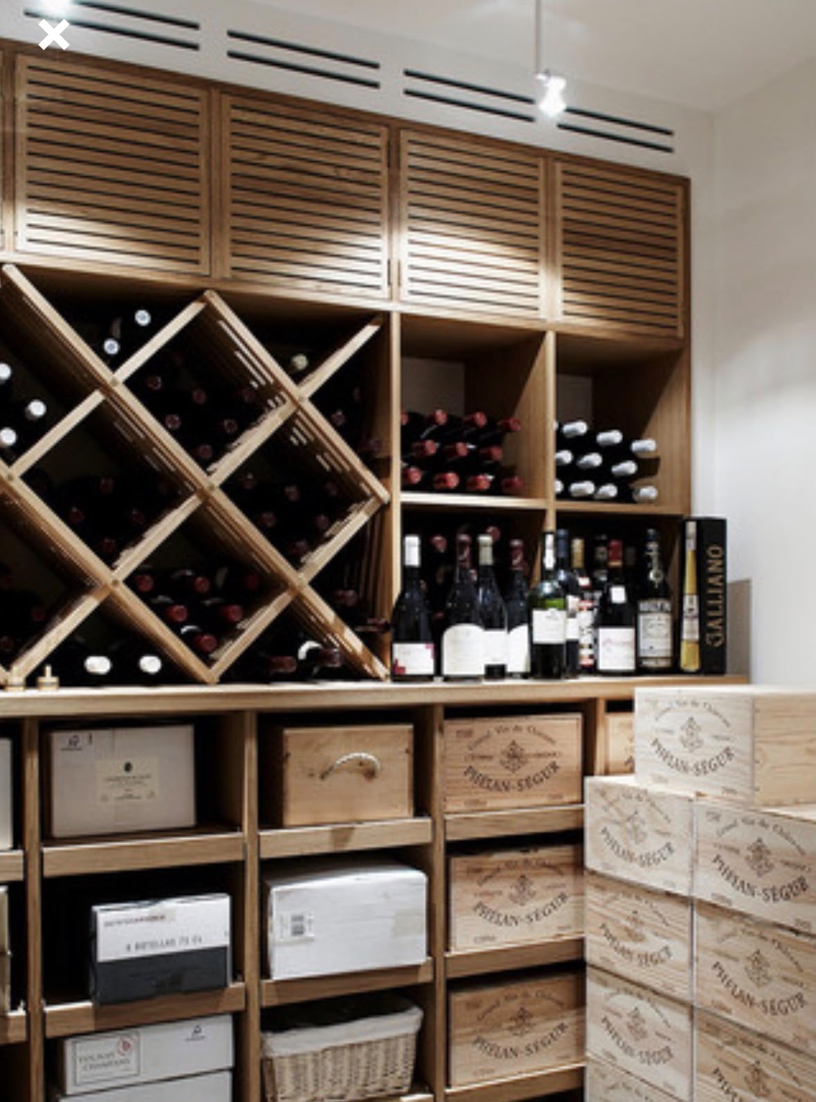 Cave à vin en bois clair avec casiers en losange, placards à claire-voie, grille de ventilation au plafond et caisses de vin empilées