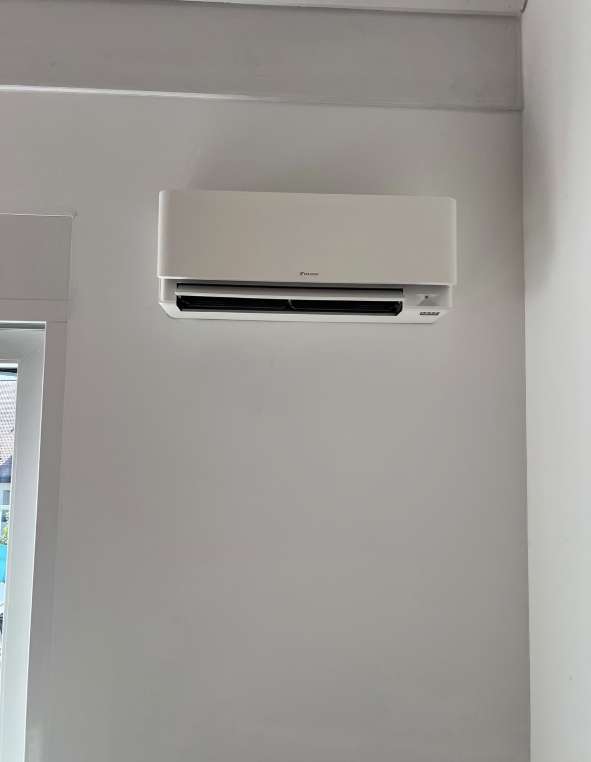 Unité murale de climatisation Daikin blanche installée en hauteur sur un mur clair, près d'une baie vitrée
