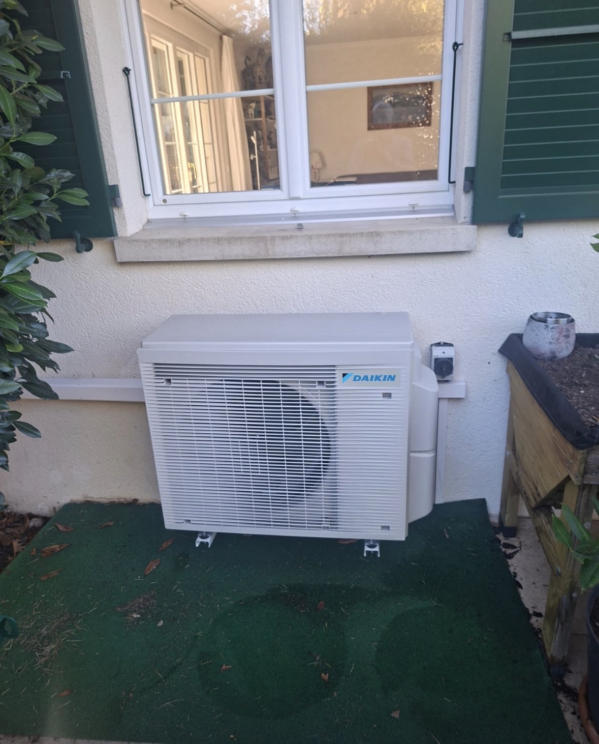 Groupe extérieur de climatisation Daikin posé sur plots au pied d'une façade blanche, sous une fenêtre à volets verts