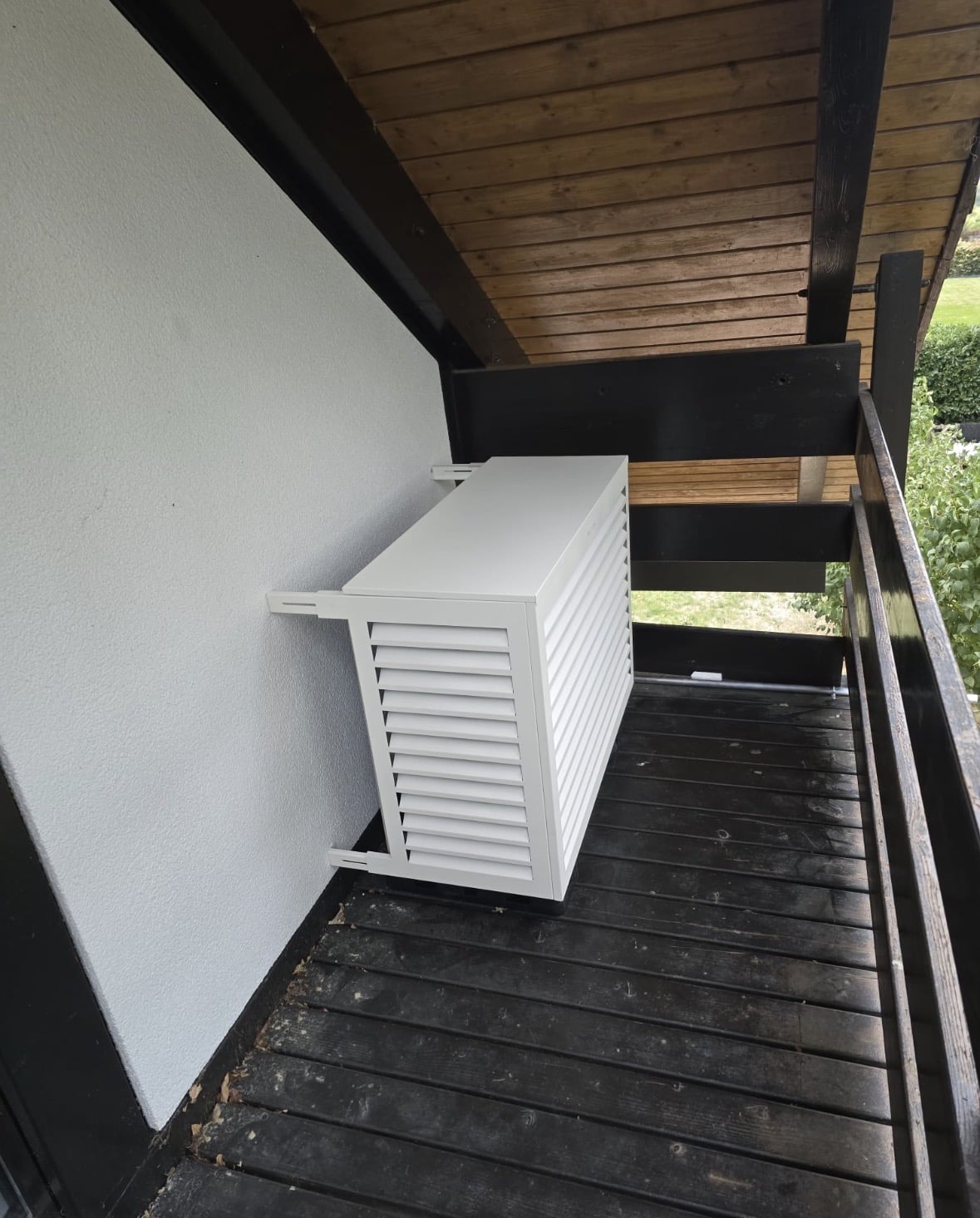 Cache Decoclim blanc à lamelles habillant l'unité extérieure de climatisation sur un balcon en bois sombre