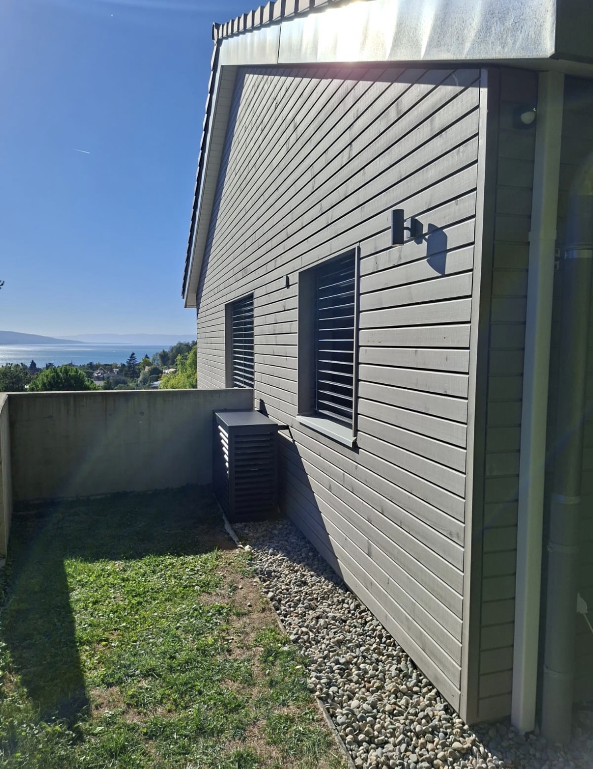 Façade en bois clair avec le groupe extérieur masqué par un capot anthracite, vue sur le lac en arrière-plan