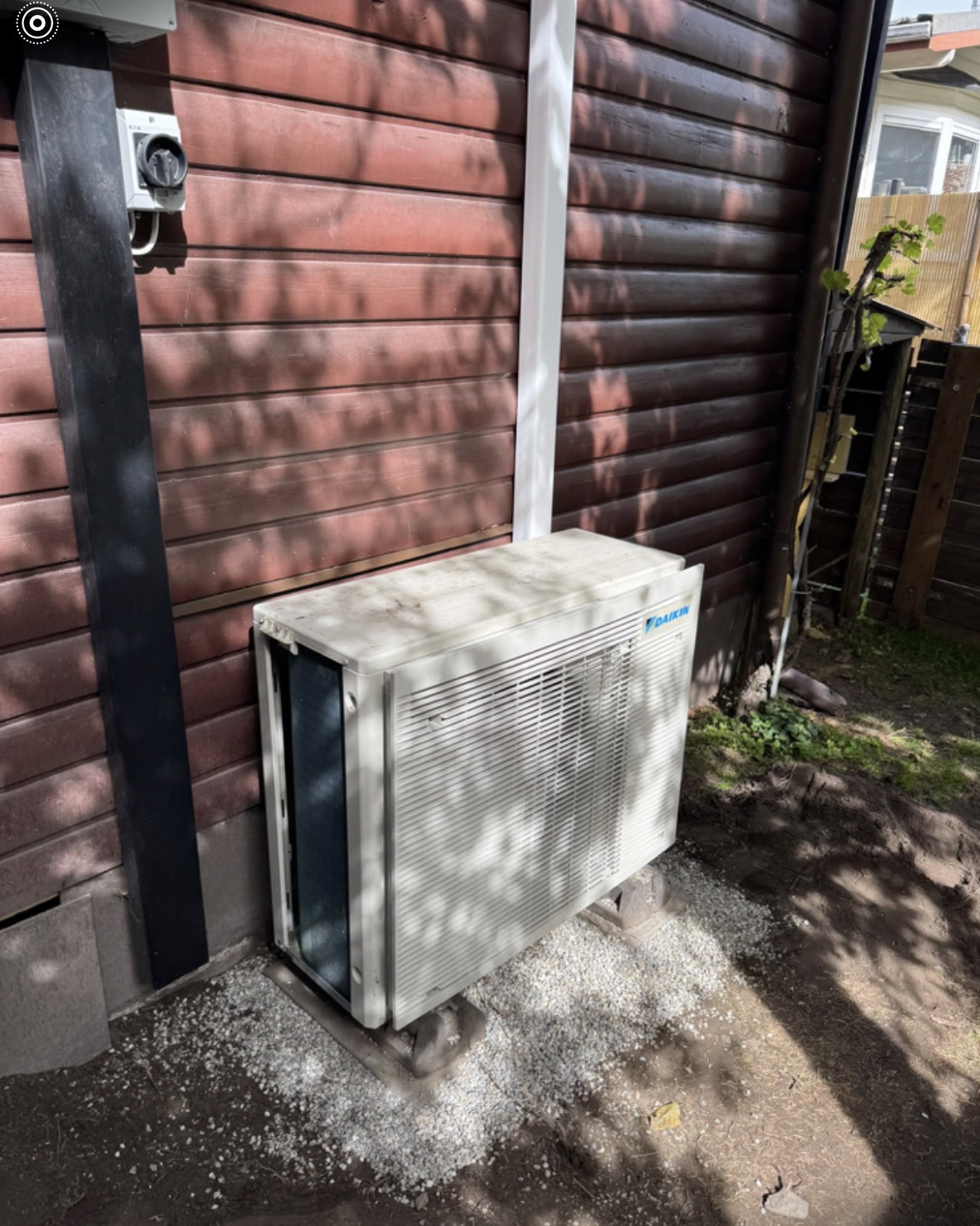 Unité extérieure Daikin posée sur un lit de gravier le long d'une façade en bois rouge d'un bungalow