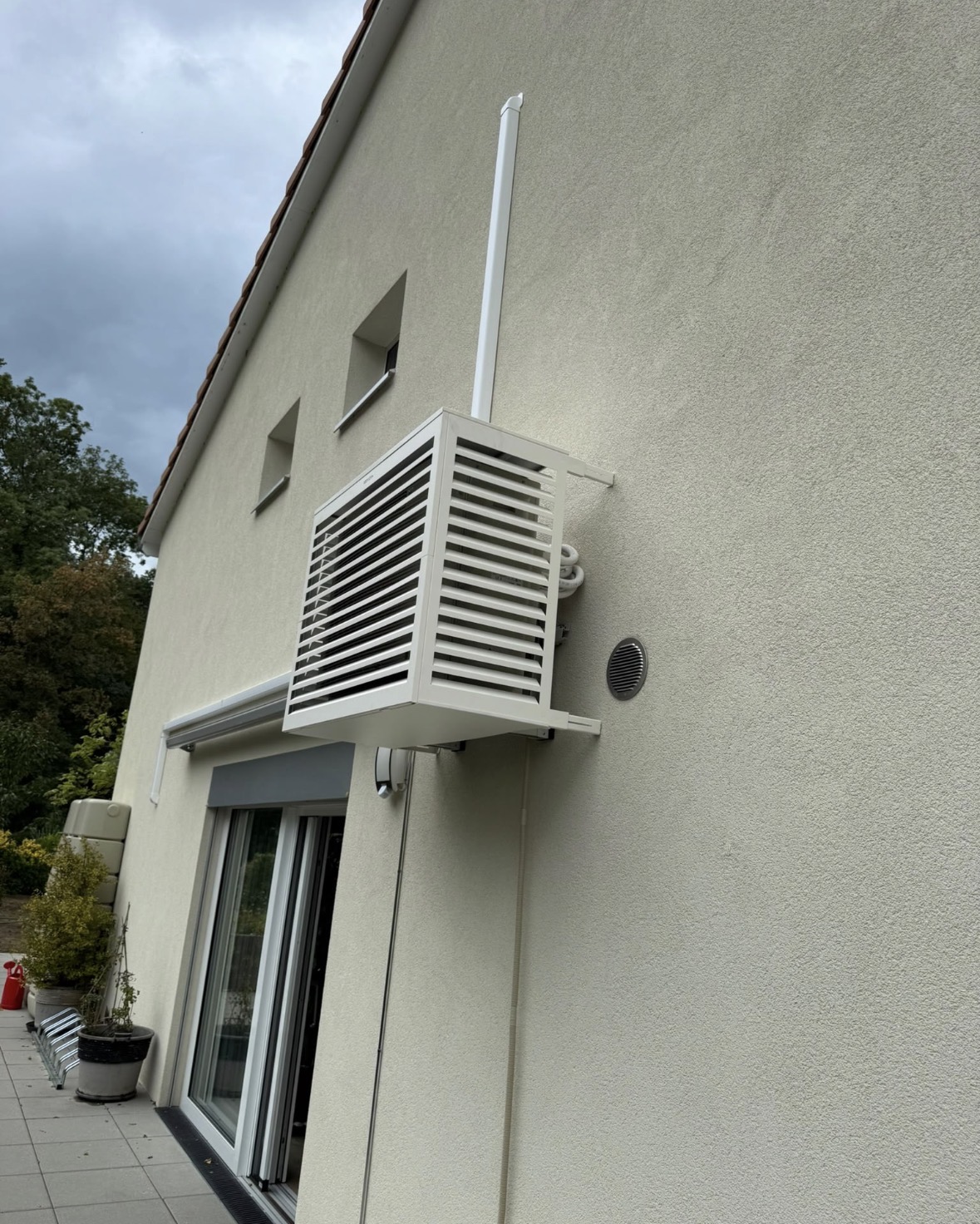 Unité extérieure de climatisation habillée d'un cache Decoclim blanc à lames, fixée en hauteur sur la façade crépie d'une villa