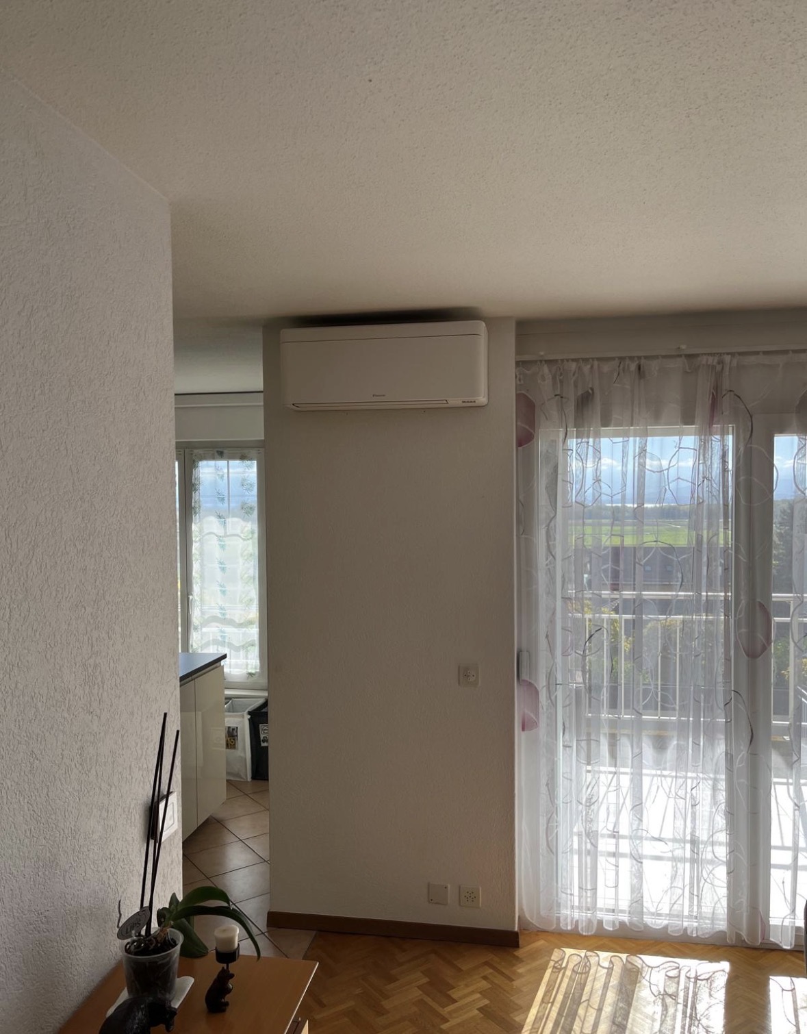 Unité murale de climatisation Daikin blanche installée en hauteur dans le séjour d'un appartement, près d'une porte-fenêtre