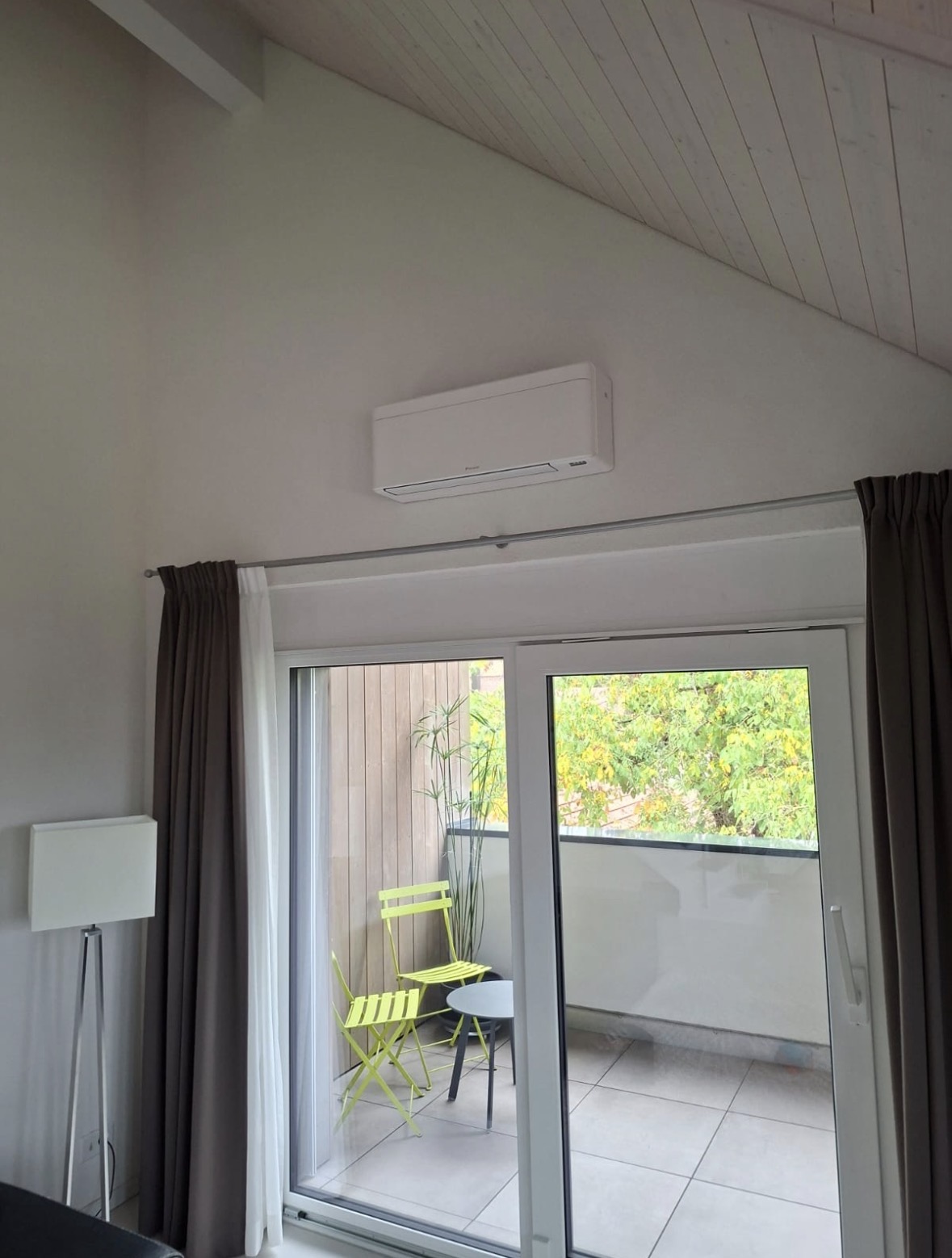 Unité murale Daikin blanche posée en hauteur près d'une baie vitrée, sous un plafond en lambris clair