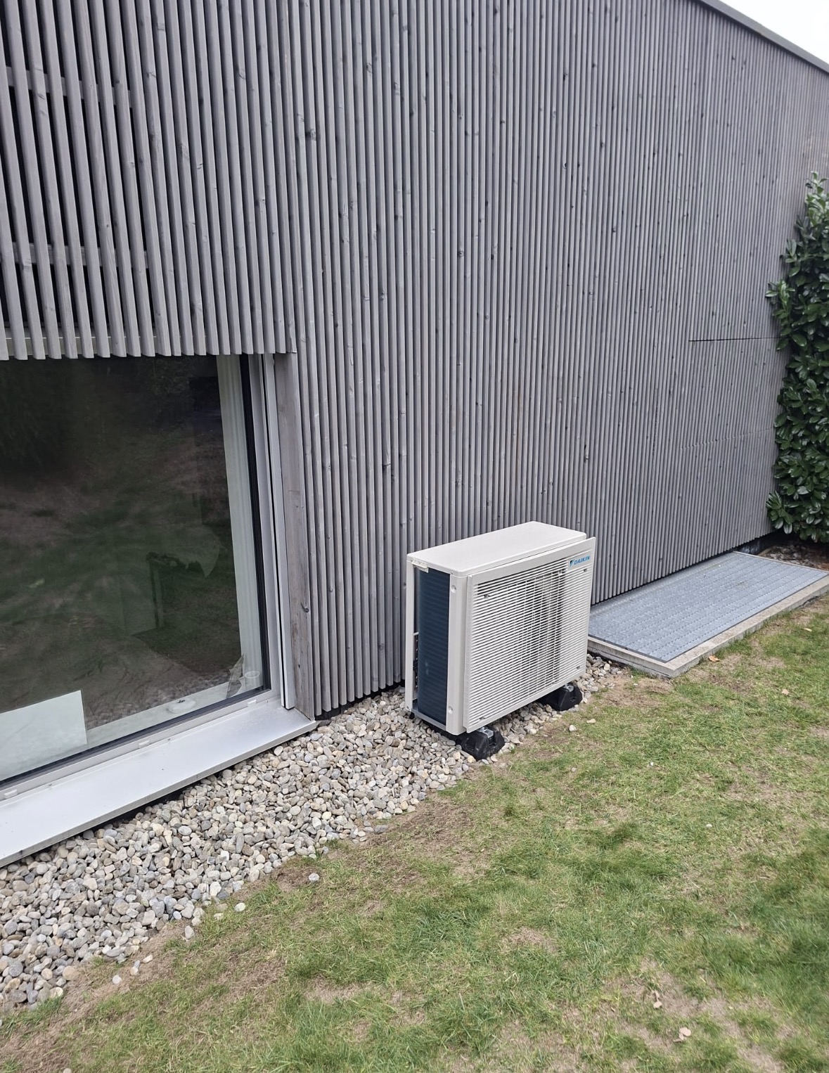 Groupe extérieur Daikin posé sur plots dans un lit de galets, le long d'une façade en bardage bois