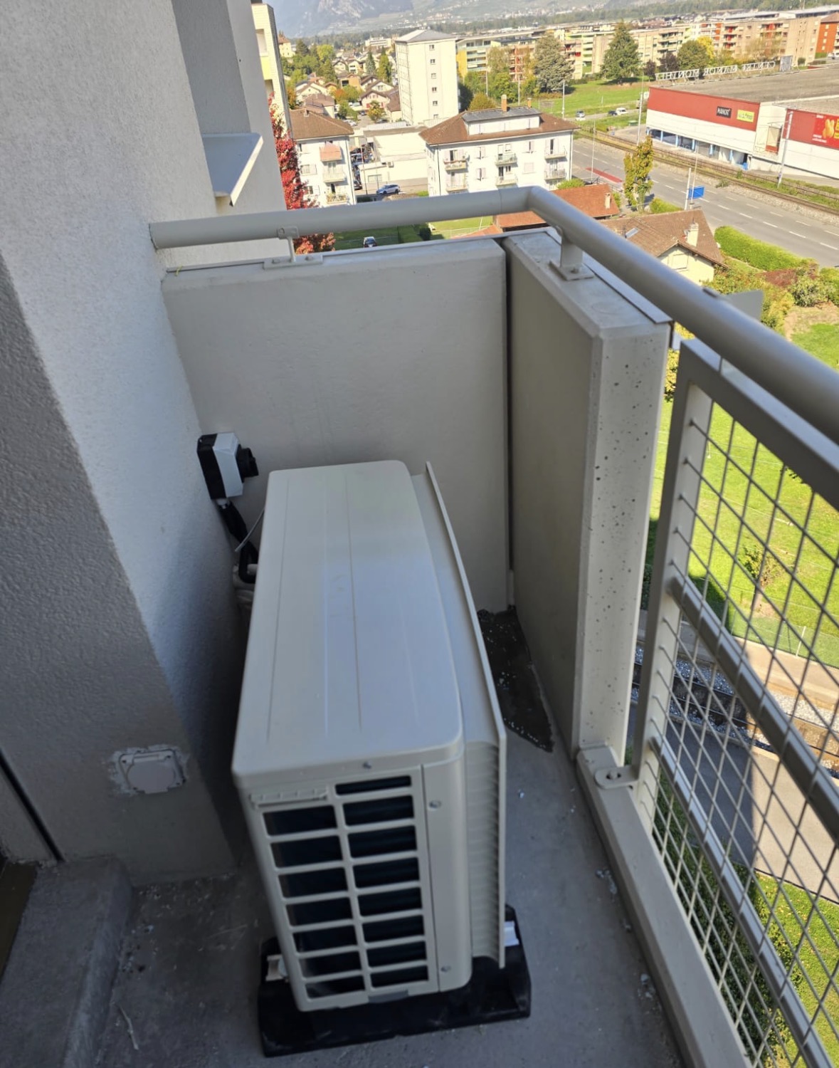 Unité extérieure de climatisation posée sur plots antivibratiles dans l'angle d'un balcon, derrière un panneau de séparation