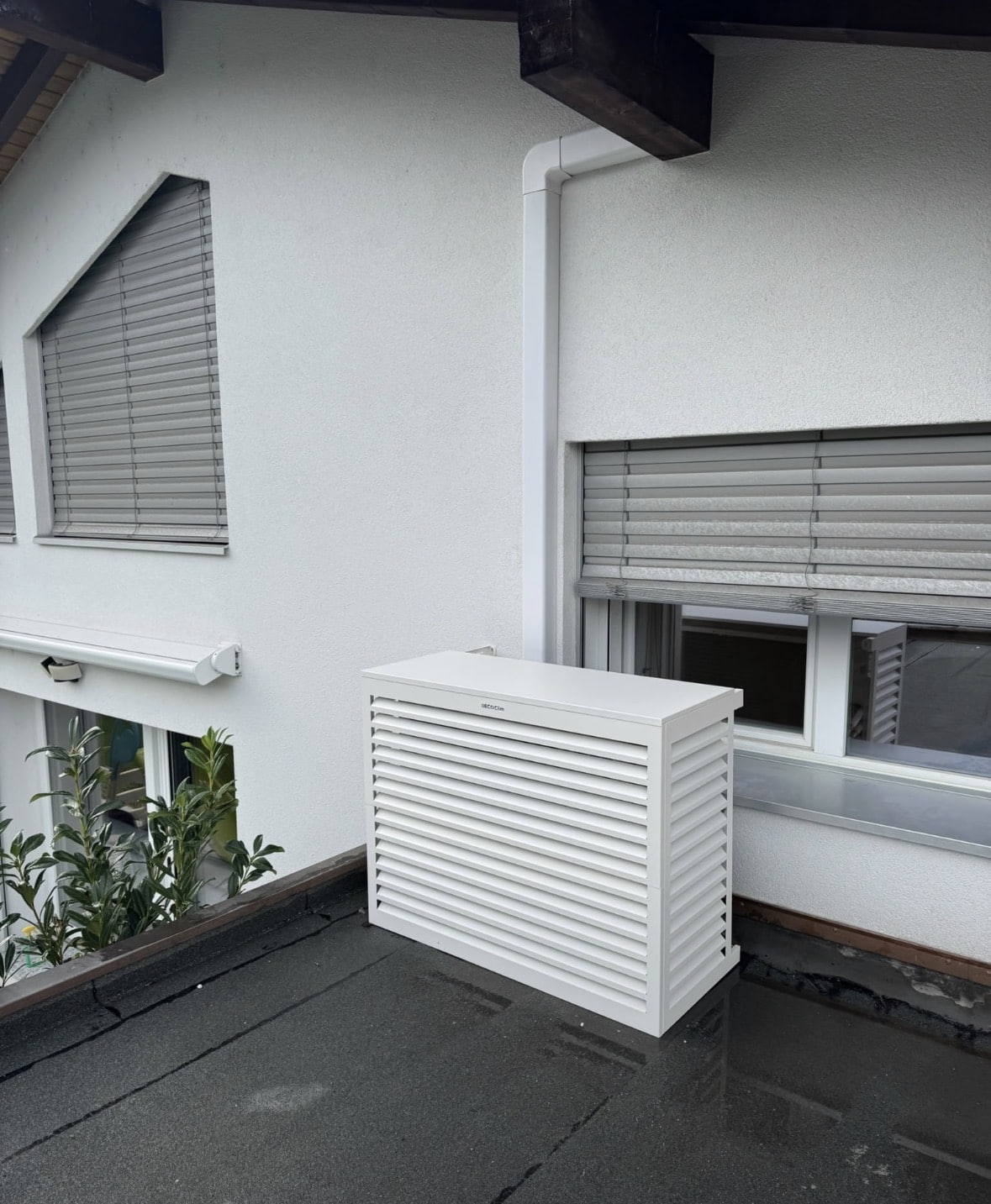 Unité extérieure de climatisation dissimulée sous un cache blanc à lamelles sur une terrasse plate
