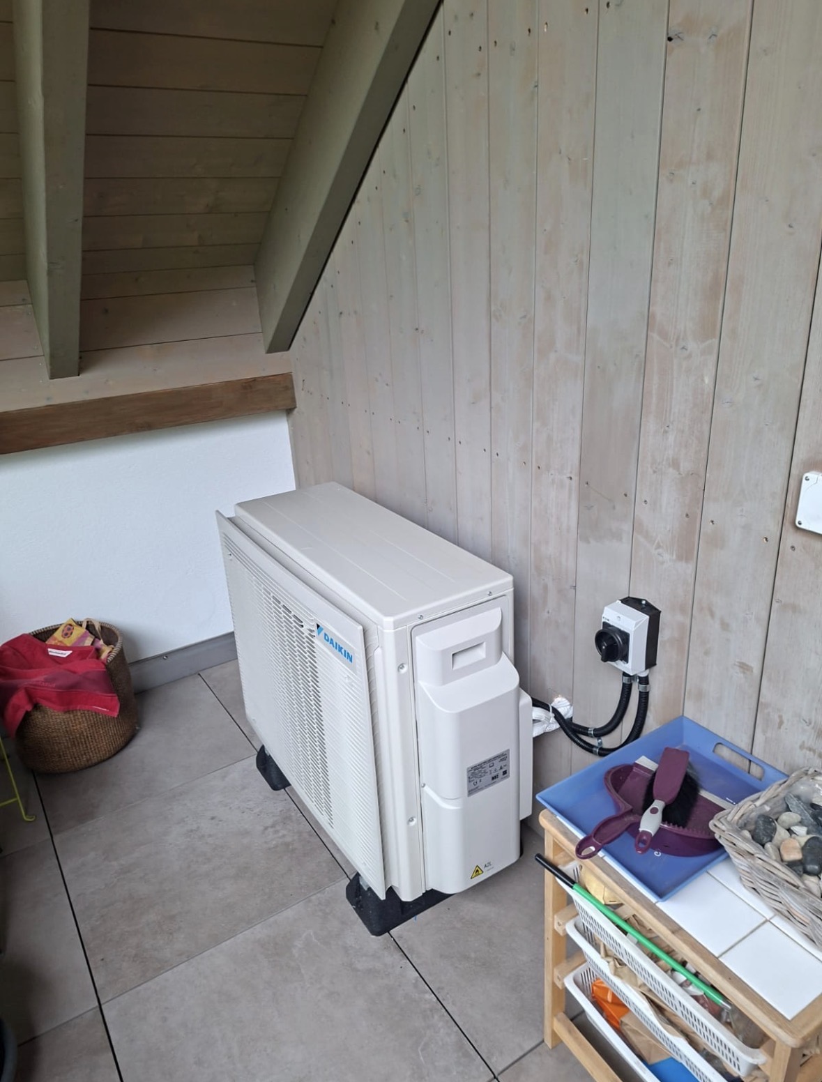 Groupe extérieur Daikin blanc sur plots, raccordé à un interrupteur de proximité fixé sur la paroi en bois