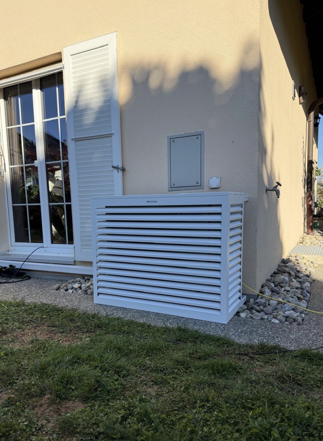 Coffrage décoratif blanc à lamelles masquant l'unité extérieure de climatisation contre une façade beige
